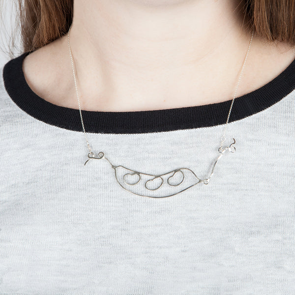 Model wears the Bean Pod Necklace, a symbolic pendant with an outline of a peapod with three beans on the inside. Sentimental jewelry that helps you celebrate all the people in your pod by Tinker Company.