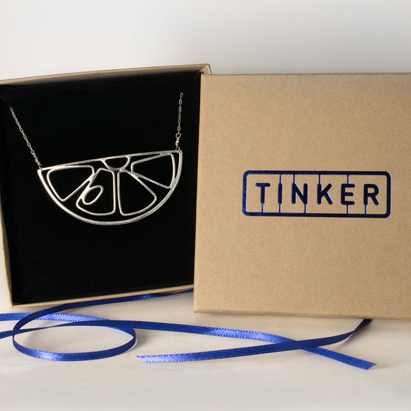 Citrus fruit pendant necklace in sterling silver with a simple minimal outline of a lemon wedge or orange slice. Part of collection of playful storytelling jewelry by Tinker Company, shown here in a Tinker gift box. 