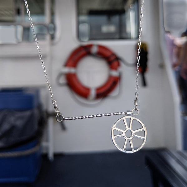 Moving Life Preserver on Rope Necklace has a lifesaver charm that moves across a twisted wire bar necklace designed to look like rope. Shown here on a boat hanging in front of a ring buoy, the inspiration for this piece of fun kinetic jewelry.
